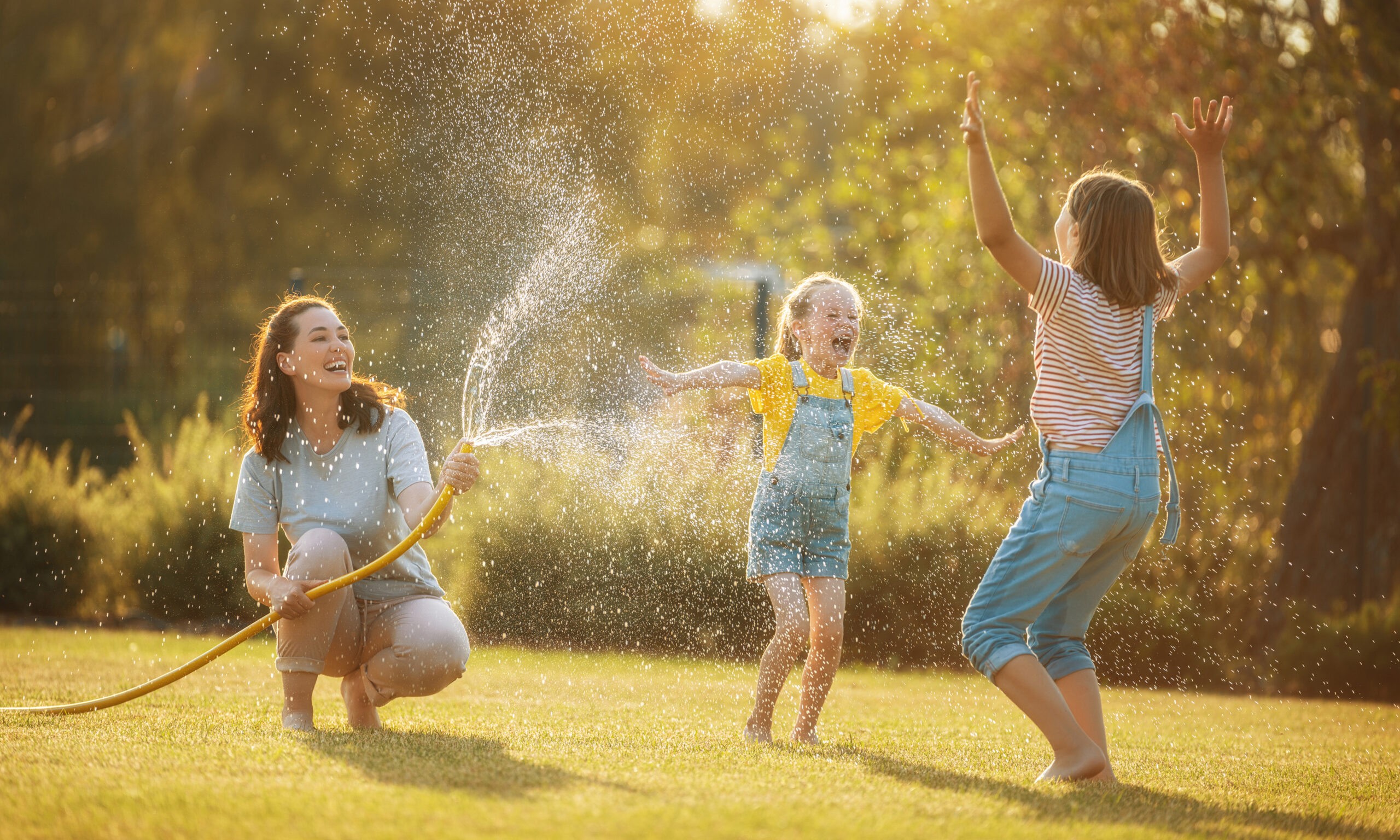 Children playing outside in the sunshine with water spray, showing fun ways families can keep kids active and healthy during summer break.